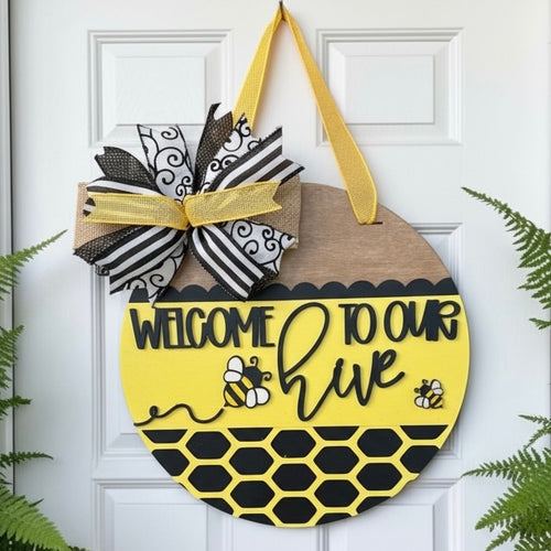 A round wooden hanging sign with a yellow and black bee-themed design that reads 'WELCOME TO OUR hive,' decorated with a black-and-white ribbon bow and gold hanging ribbon, shown on a white front door with greenery at the sides.