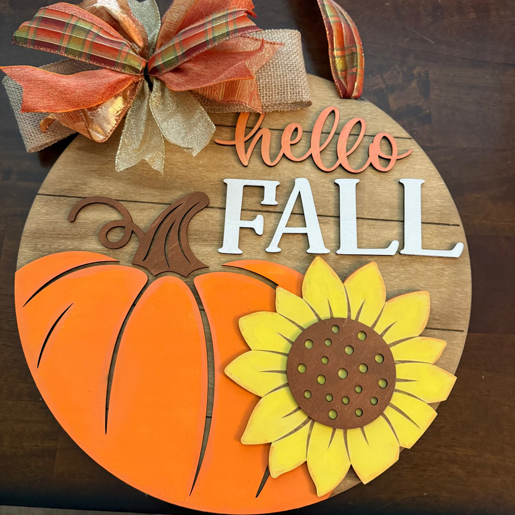 Decorative wooden sign with 'hello FALL' text, pumpkin, sunflower, and plaid bow on a dark background.